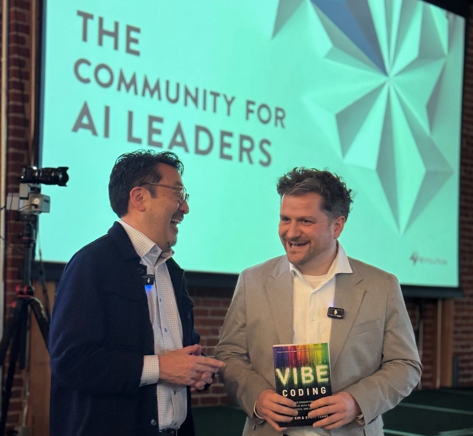 Gene Kim laughing on stage in Munich next to a guest holding a copy of Vibe Coding, in front of a screen reading “The Community for AI Leaders”